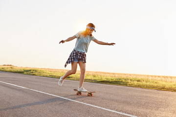 Fototapeta premium Young attractive female skater riding outdoor on asphalt road, raised her arms, sporty woman wearing casual clothing skateboarding alone in sunset in summer.