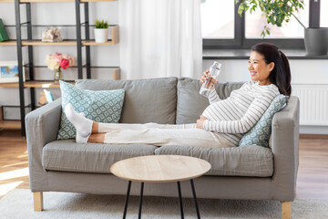 pregnancy, rest, people and expectation concept - happy smiling pregnant asian woman sitting on sofa at home and drinking water from reusable glass bottle