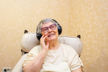 A cute elderly lady with a dreamy smile sits in an armchair and listens to music or an audiobook on...