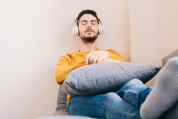 Young adult man 30s relaxed, comfortable and calm, listening to music with his wireless headphones indoors. Therapy, music and relaxation concept.