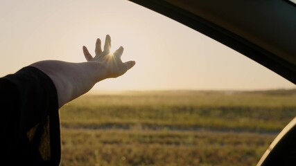 A free woman drives a car, catches sunrise light and wind with her hand from car window. Vacation, travel. Girl sits in front seat of car, reaches out to window and catches glare of setting sun.