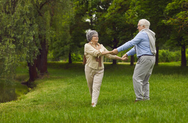 Naklejka na ściany i meble Happy elderly pensioners, man and woman, husband and wife, dancing together in the park on the green grass, breathing fresh air, useful walks.