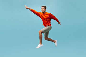 Fototapeta na wymiar Positive guy with beard in bright cool clothes and white modern sneakers looking away and jumping on isolated blue background. .