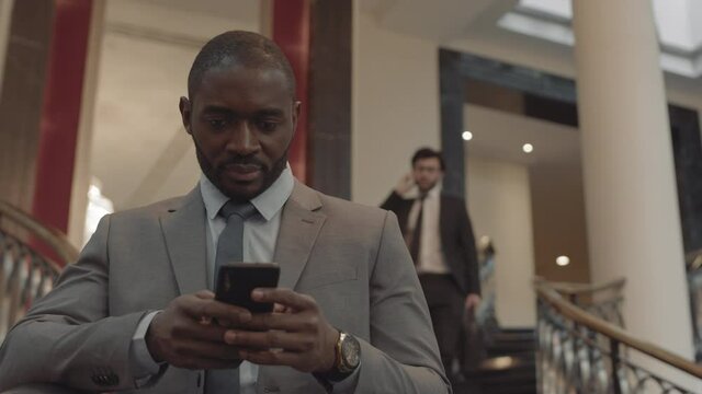 Chest-up of confident African manager wearing grey suit, using smartphone, standing in stairwell of luxurious building, blurred colleague going down stairs on background