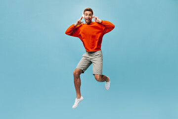 Obraz premium Handsome guy in good mood in orange sweatshirt, shorts and white sneakers looking into camera, listening to music and jumping..