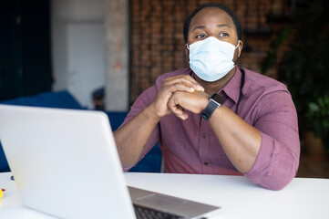 Pensive African-American guy wearing medical mask looks away sitting at the desk in front of laptop, a black male entrepreneur worried about crisis in business due to quarantine