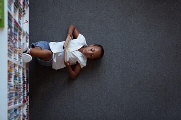 Overhead view of african american schoolgirl lying on floor reading book in school library