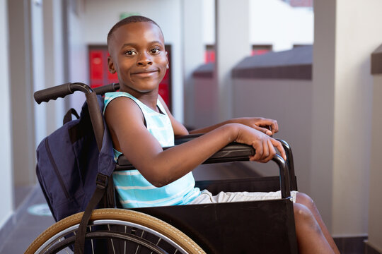 Portrait of smiling disabled african american schoolboy sitting in wheelchair in school corridor