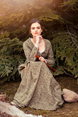 Fototapeta premium Portrait of Calm Caucasian Female in Old Medieval Green Dress With Flute Against Firtrees Outdoors.