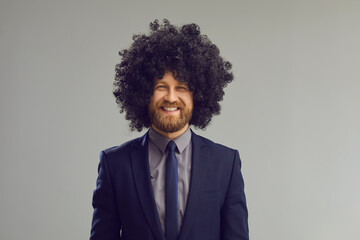Obraz premium Headshot of cheerful smiling young man with crazy curly hair. Portrait of happy positive stylish bearded businessman in suit, necktie and funny wig looking at camera standing on grey studio background