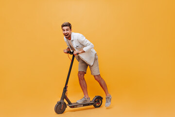 Funny fashionable cool guy in denim shirt and grey sneakers riding scooter and having fun on isolated orange background.. © Look!