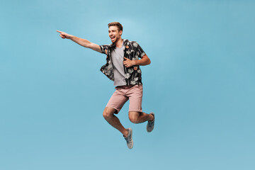 Fototapeta na wymiar Cool fashionable man with beard in grey t-shirt, printed shirt and pink shorts looking away, rejoices and jumping on blue backdrop..