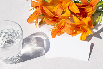 Mockup of business cards, a glass of water and a bouquet of orange lilies on a white table. Hard...