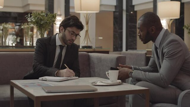 Medium long of dark-haired manager wearing black suit, interviewing male African candidate in grey formalwear, sitting at coffee table in lobby