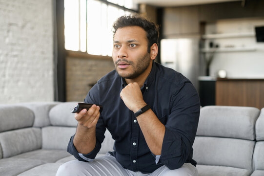 Breaking News Concept. Amazed Indian Guy Sits On The Couch With A Remote Controller In Hand, Concerned Face Expression, Watching TV At Home