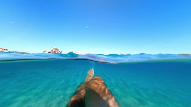 Split-shot, stunning view of a tourist swimming on a beautiful and turquoise water. Caprera, La Maddalena Archipelago, Sardinia, Italy. Over-under shot, half underwater, half sky.