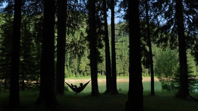 Young man sitting in a hammock and enjoying a beer. The shore of the mountain lake in the green forest. Green mountain forest lake in summer season. Calm place to bee. Chilling in the woods