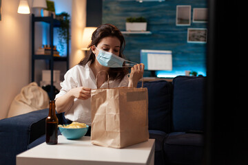 Cheerful woman taking medical protection face mask out after purchasing takeaway food in evening. Smiling caucasian female enjoying delicious junk-food home delivered. Tasting box order