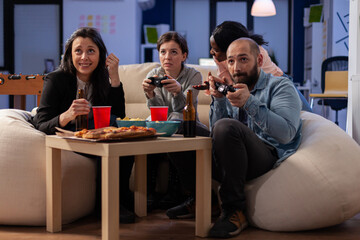 Diverse team of workers playing console game on tv at office after work using joysticks on sofa. Multi ethnic friends starting competition while having snacks pizza chips and drinking beer