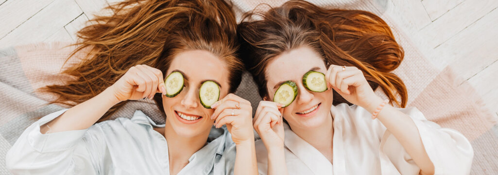 Two girls make homemade face and hair beauty masks. Cucumbers for the freshness of the skin around the eyes. Women take care of youthful skin. Girlfriends laugh at home lying on the floor on pillows.