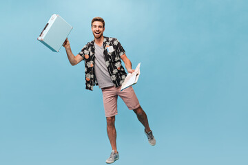 Joyful young man with ginger beard and stylish hairstyle in grey t-shirt and shirt sleeve shirt and sneakers holding suitcase and map..