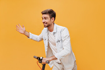 Joyful trendy man with ginger beard in light t-shirt and denim jacket smiling, looking away and posing with scooter on orange backdrop..