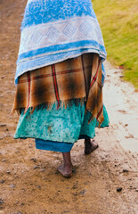 A barefoot woman covered with multiple blankets walking barefoot on a wet muddy road