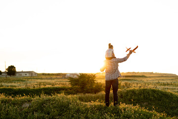 Family single father with his daughter at sunset in a field on a summer evening launching a toy airplane into the air. baby girl sitting on dad's shoulders looks the sky.childhood concept
