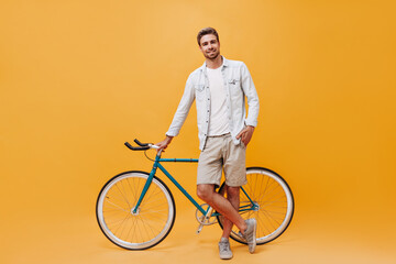 Modern cool young man with brown hair and red beard in white t-shirt, denim jacket and stylish sneakers posing with bicycle..