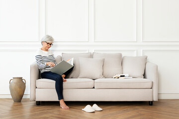 Senior woman reading a book on the sofa in a Scandinavian decor living room - Powered by Adobe