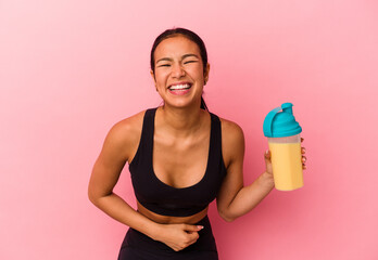 Young Venezuelan woman drinking a protein shake isolated on pink background laughing and having fun.
