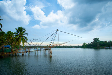 A Chinese fishing net and a small hut with coconut trees under a blue and white cloudy sky, River photography with a Chinese fishing net from Kerala, India