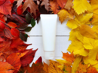 White squeeze bottle cosmetic tube and round frame of bright autumn or fall leaves on white wooden...
