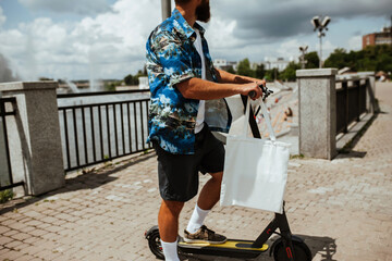 Young hipster man driving an electric scooter on a bright sunny day holding white textile eco bag...