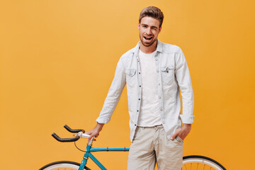 Fototapeta na wymiar Cool trendy man with brown hair in stylish light clothes winking, smiling and posing with bike on isolated orange background..