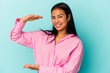 Young Venezuelan woman isolated on blue background holding something little with forefingers, smiling and confident.