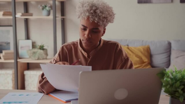 African American female student secretary freelancer business woman sitting at home office table studying looking at laptop reads reports compares data on paper and on computer screen analyzes project