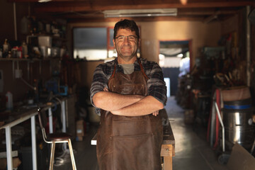 Smiling caucasian male knife maker with crossed hands in workshop looking at camera