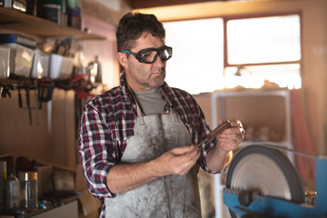 Caucasian male knife maker wearing apron and glasses , making knife in workshop
