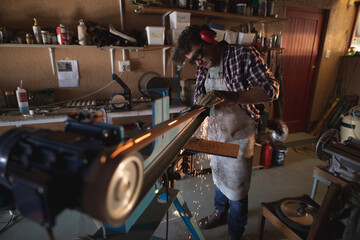 Caucasian male knife maker wearing apron and glasses, using sander in workshop