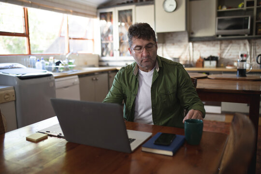Caucasian man in kitchen sitting at table using laptop and writing notes