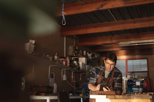 Caucasian male knife maker sitting at desk, using tablet in workshop