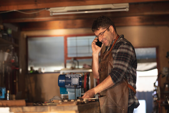 Caucasian male knife maker standing at desk, talking by smartphone in workshop