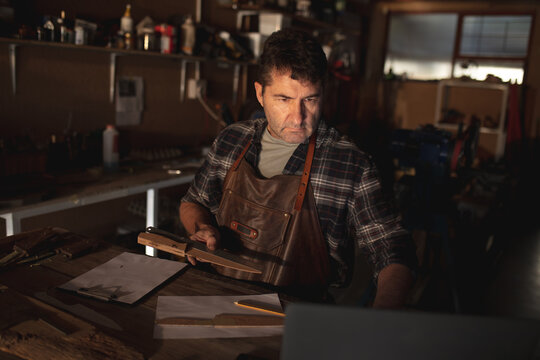 Caucasian male knife maker holding knife, using laptop in workshop