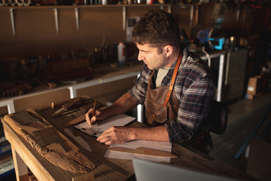 Caucasian male knife maker sitting at desk, making notes in workshop
