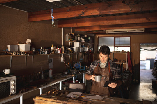 Caucasian male knife maker standing at desk, drinking coffee, using tablet in workshop