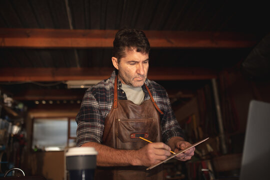 Caucasian male knife maker sitting at desk, making notes in workshop
