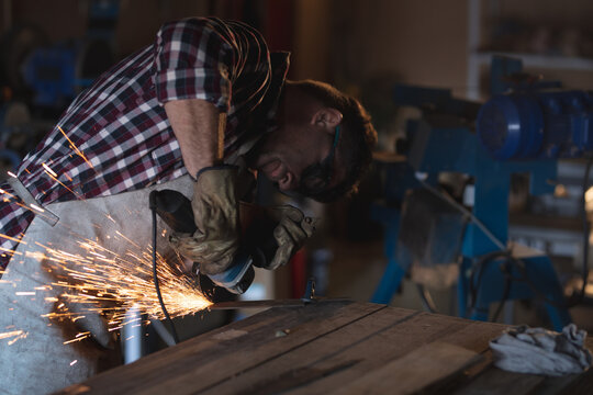 Caucasian male knife maker wearing apron and glasses, using angle grinder in workshop - Powered by Adobe