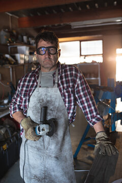 Caucasian male knife maker wearing apron and glasses, holding angle grinder in workshop