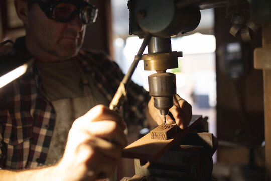 Caucasian male knife maker wearing apron and glasses, using drill in workshop